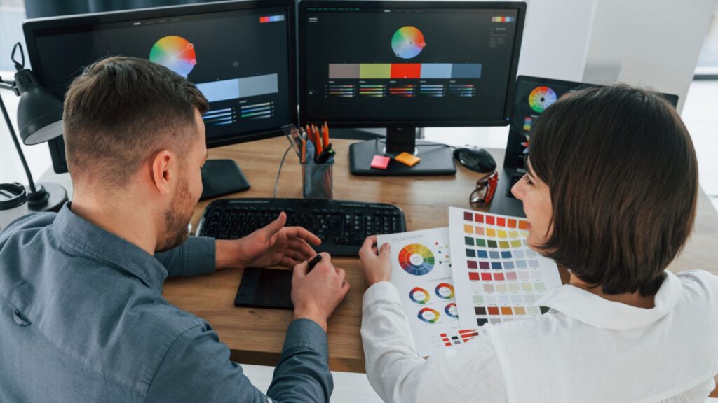 Two people sit at a desk with color swatches and color wheels on monitors, collaborating on graphic design and planning an explain services animation.