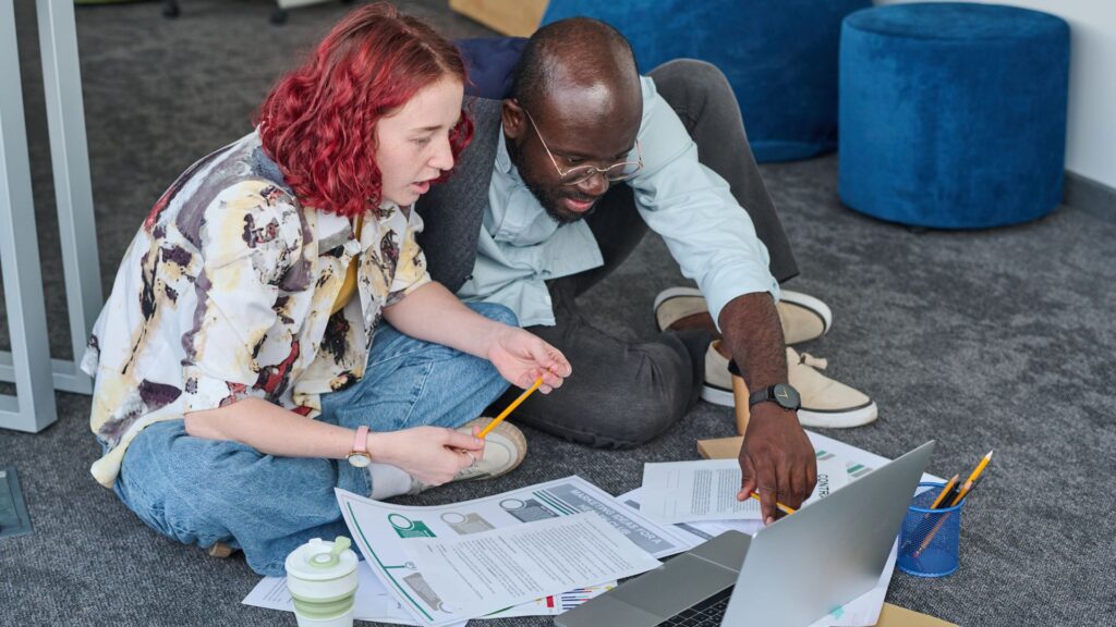 Two people sitting on the floor surrounded by papers, discussing documents and looking at a laptop screen together while reviewing an animation value guide and exploring 30 second animation cost options.