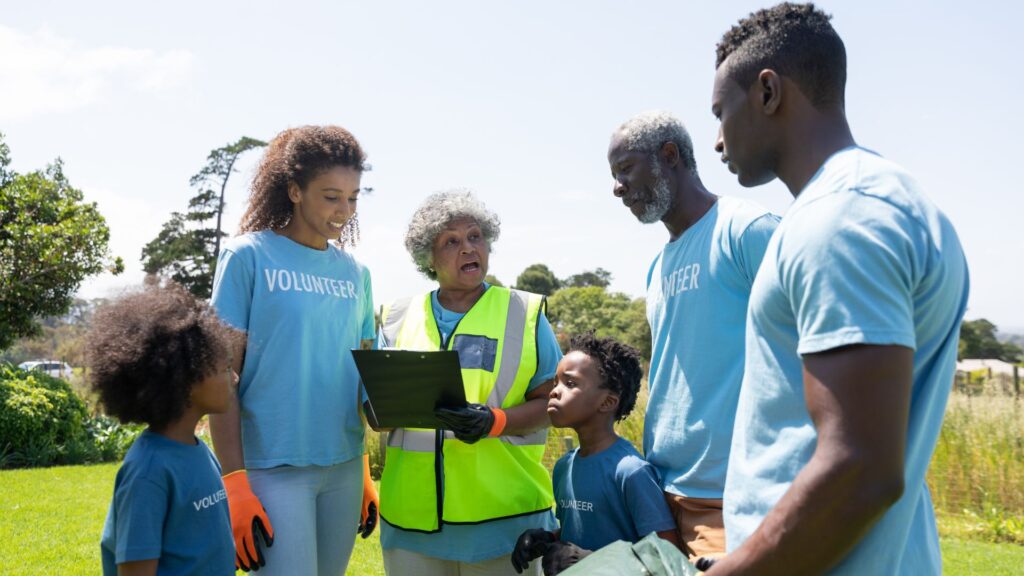 A group of volunteers, including adults and children, stand outdoors in a park, listening to a woman in a safety vest holding a clipboard as she discusses upcoming projects funded by Nonprofit Animation Grants.