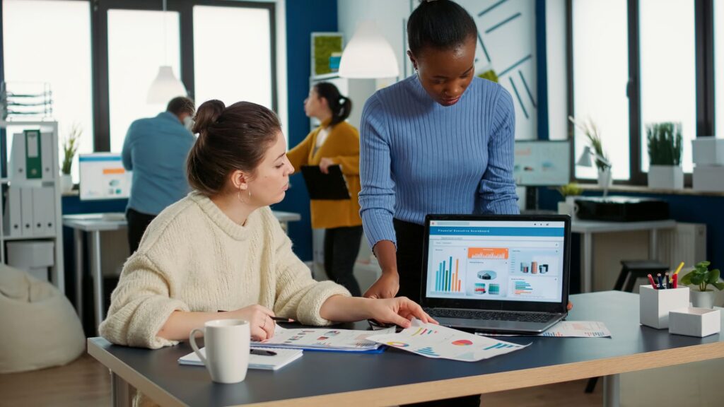Two women in an office review printed business charts and graphs, with additional data and impactful animation displayed on a laptop screen. Other colleagues work in the background.