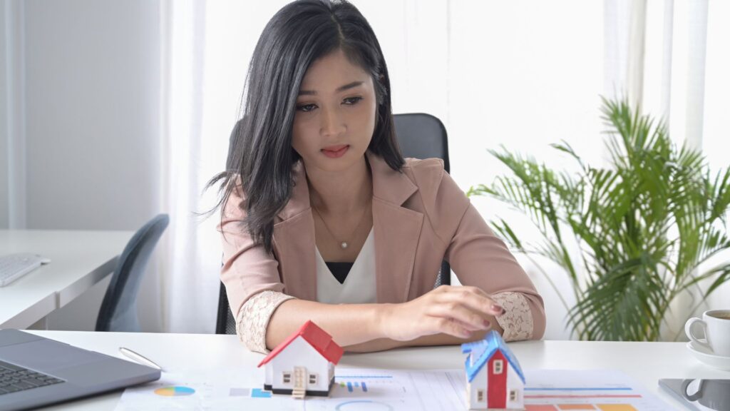 A woman sits at a desk, moving a model house on a document with charts, visualising loans. Another model house and a laptop are also on the desk.