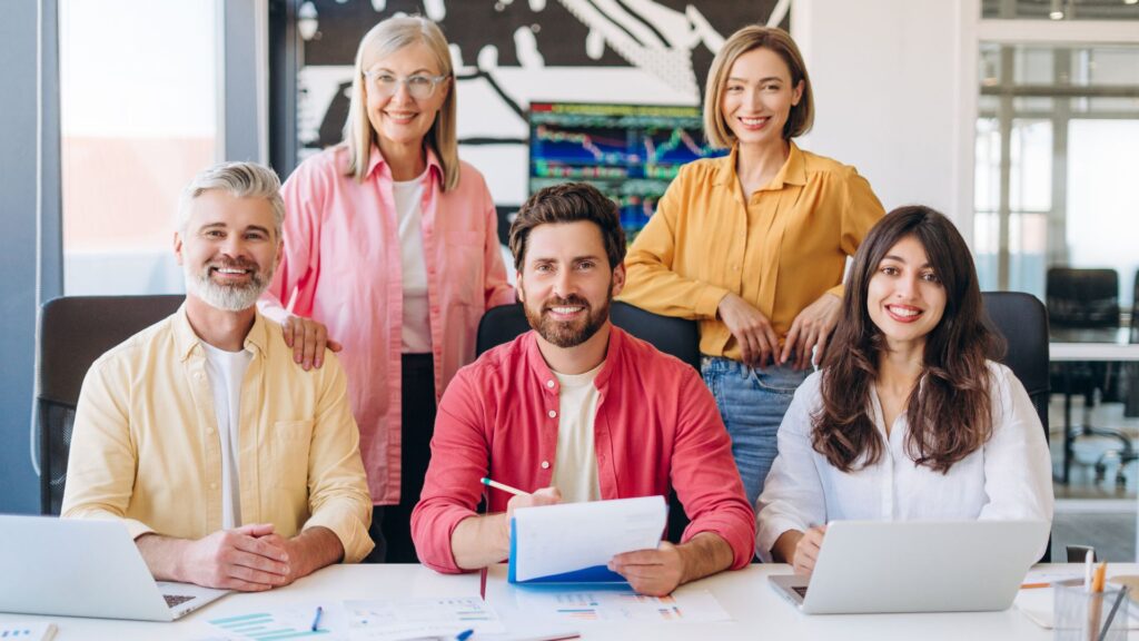 Five people smiling and posing together at a desk with laptops and documents in a modern office setting, brainstorming ideas for an engaging onboard customers animation.