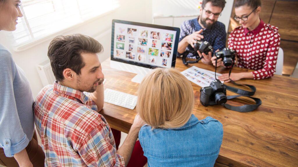 Four people collaborate at a wooden desk with cameras, photo prints, and a computer displaying photo thumbnails—brainstorming ideas for an engaging animation for landing page concepts.
