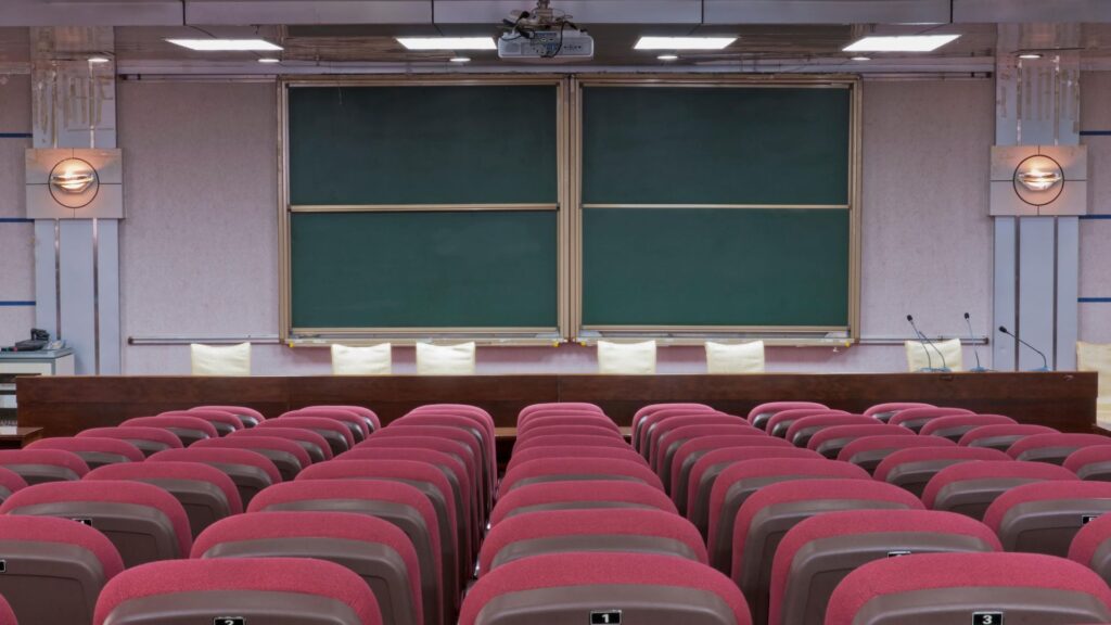 Rows of empty red chairs face a podium and large double green chalkboards in a lecture hall classroom, setting the perfect scene for an engaging Educational Animation or Animation Guide at a leading educational institution.