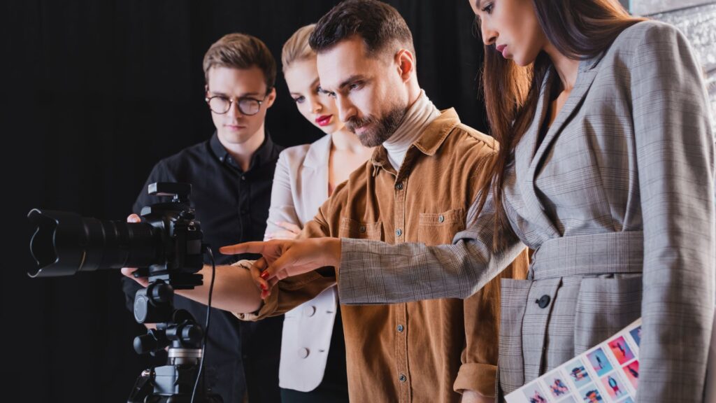 Four people stand together, closely examining a professional camera on a tripod—one points at the camera while another, from a Best Animation Company, holds a contact sheet with photo thumbnails.