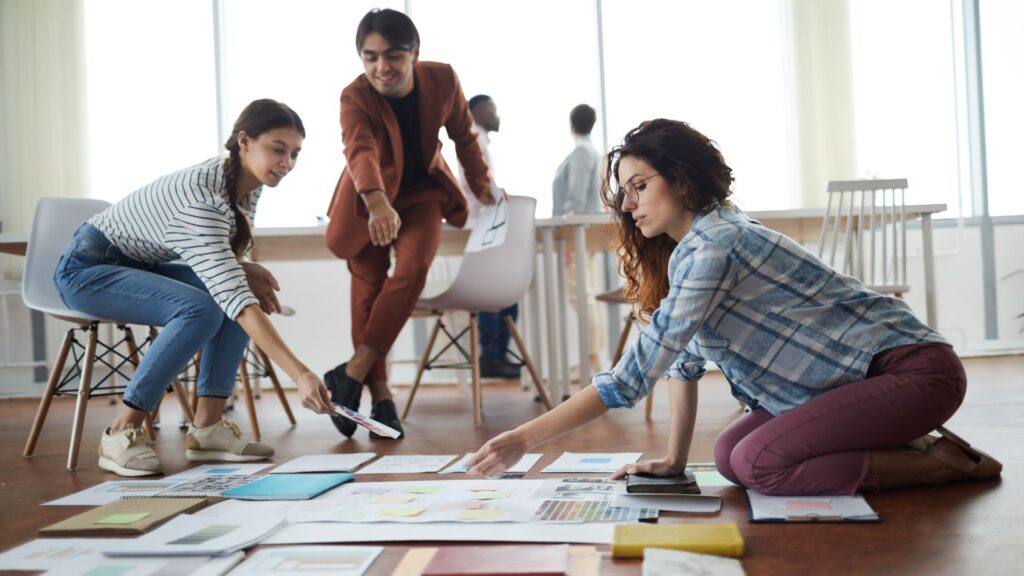 Three people collaborate on the floor, reviewing papers and documents spread out in front of them in a bright office space, visualizing ideas for a Change Management Animation project.