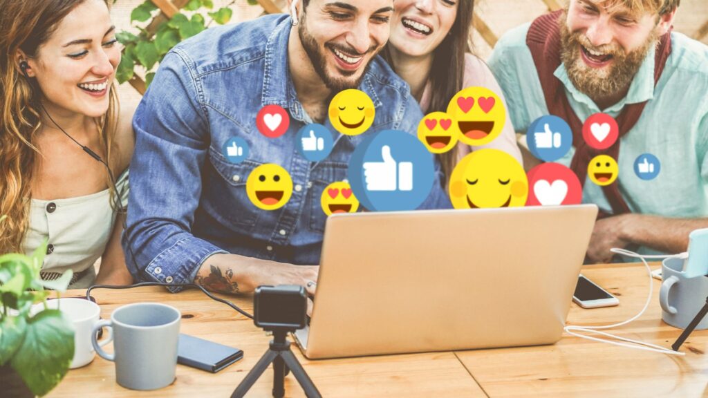 Four people smile and look at a laptop outdoors with social media emoji icons, such as thumbs up and hearts, overlaid on the image—perfect inspiration for animation for social media. Coffee cups and a tripod sit on the wooden table.