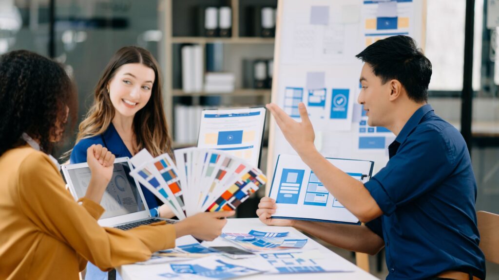 Three people sit at a table discussing design layouts and color palettes, with printed samples and a laptop, while a board with wireframes and a Change Management Animation plan is visible in the background.