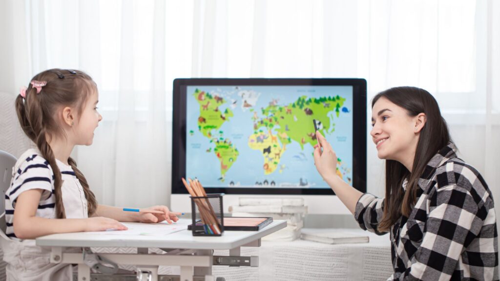 A woman and a young girl sit at a desk, looking at a world map displayed on a computer screen featuring an educational animation as the woman points to a location.