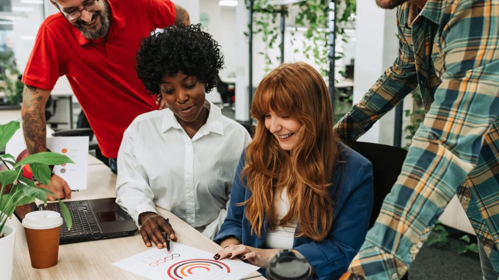 Four people gather around a desk, reviewing a printed chart with concentric circles. Two are seated, two standing, as they discuss Change Management Animation strategies beside a laptop and coffee cup on the table.