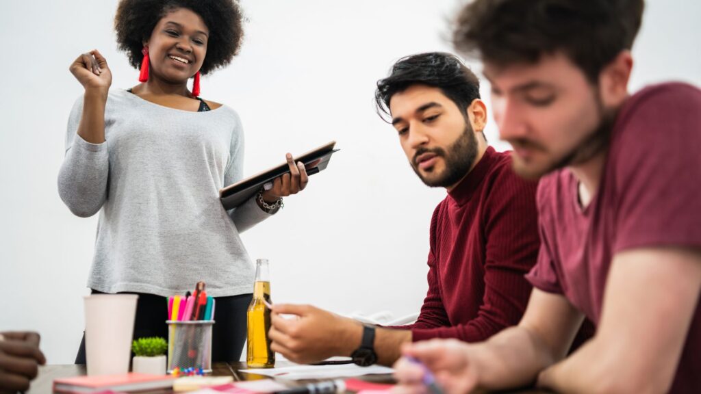 A woman stands holding a tablet and smiling while three men sit at a table with drinks, papers, and stationery, engaged in discussion about Change Management Animation.