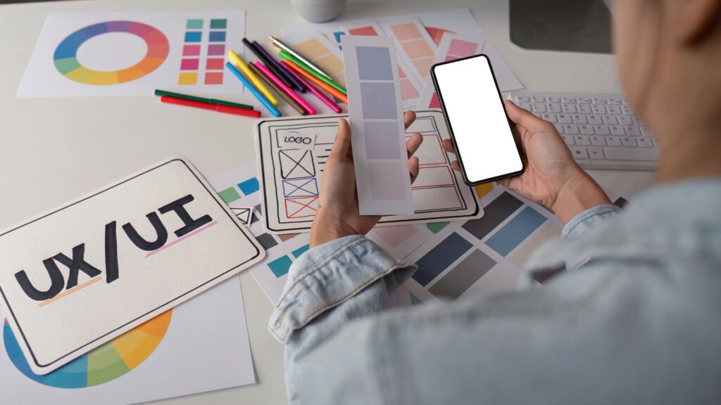 Person working at a desk with UX/UI design materials, holding a smartphone and color swatches, surrounded by color charts, pens, and a keyboard—exploring animation for mobile app interfaces.