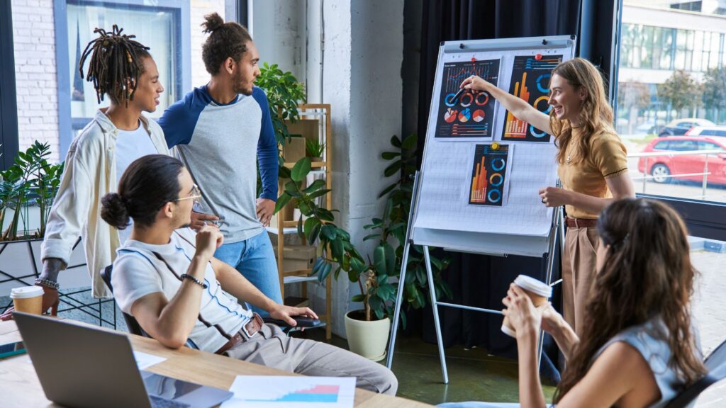Five people in a modern office discuss charts on a flipchart; one person stands and points while others listen, using laptops and coffee cups—perfect for illustrating Explain Services Animation presentations.