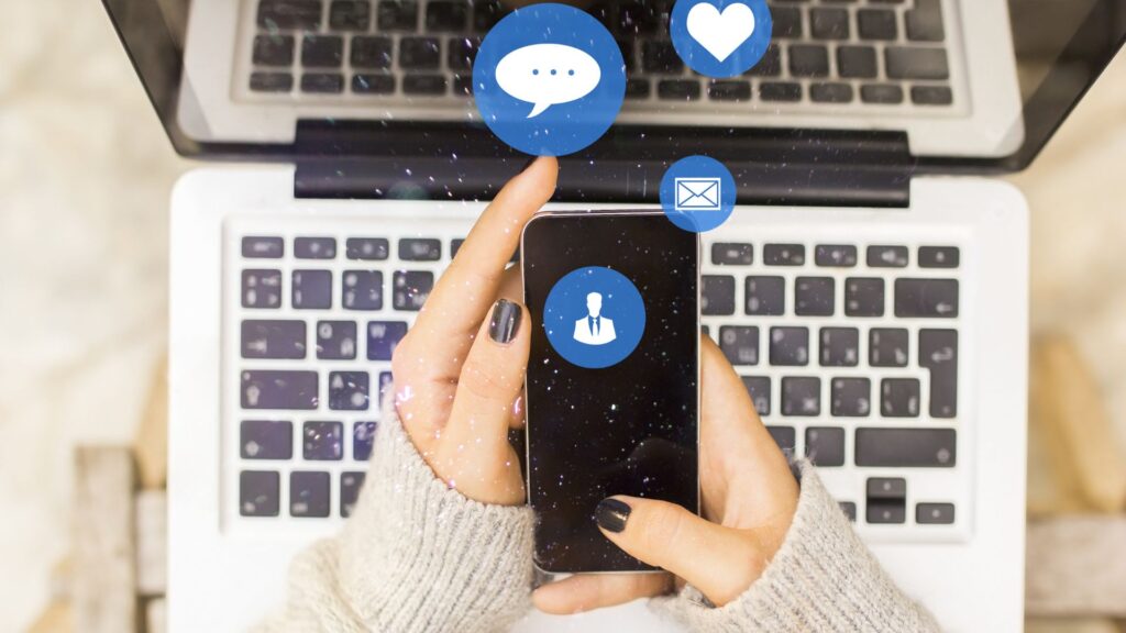 A person in a gray sweater holds a smartphone in front of an open laptop, with Animation for Social Media notification icons hovering above the phone.