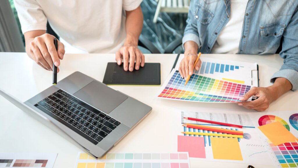 Two people sit at a desk with a laptop, tablet, color swatches, and charts, collaborating on color selections for an animation production, carefully planning every detail as part of their step-by-step guide for the project.
