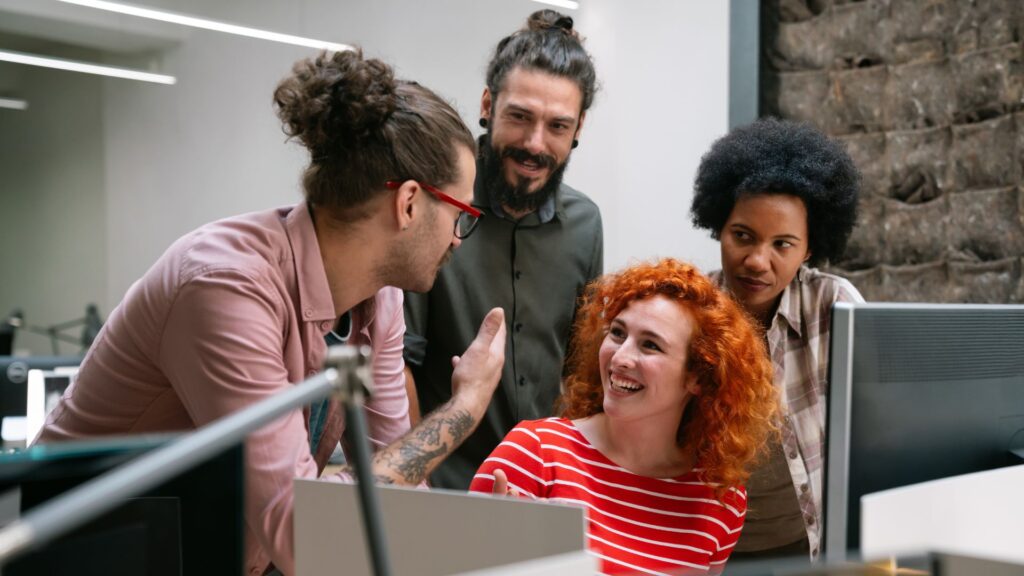 Four people gathered around an office desk, engaged in a discussion about animation production; one person is seated and smiling, while the others stand and listen attentively.