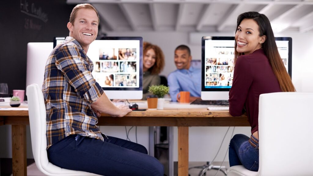 Two people sit at a desk working on computers, smiling at the camera, as they collaborate on animation production, with two more colleagues blurred in the background.