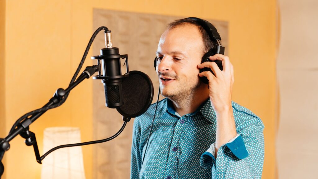 Man in a blue patterned shirt wearing headphones, standing in front of a microphone in a recording studio, preparing to record voiceover for a 2 minute animation.