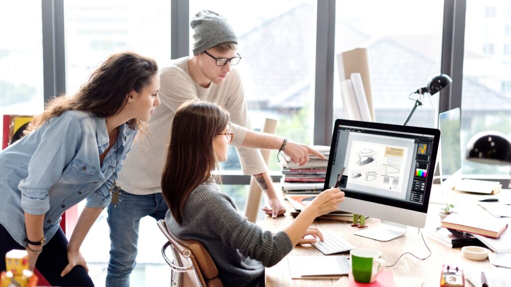 Three people collaborate at a desk, reviewing animation production sketches on a large computer monitor in a modern office with large windows.