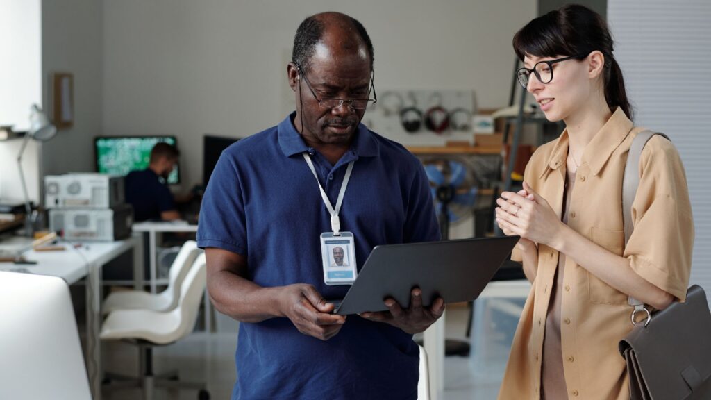 Two people stand in an office; one, wearing an ID badge, holds and looks at a laptop showcasing a Tech Product Launch Animation. The other speaks and gestures with a bag. Office equipment and another person appear in the background.