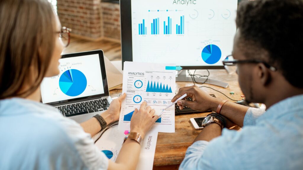 Two people reviewing printed analytic charts at a desk, with additional charts—including a Tech Product Launch Animation—displayed on a laptop and monitor in the background.