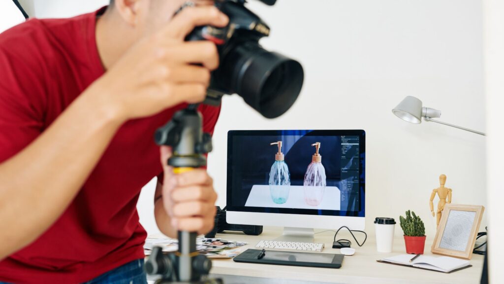 A person in a red shirt photographs two soap dispensers, which are displayed on a computer screen in a home office setting, possibly researching explainer video price trends in the UK or 2025 cost estimates.
