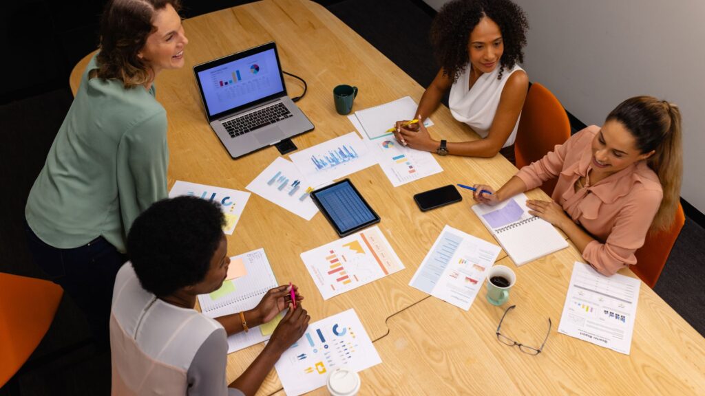 Four women sit around a table in a UK meeting room with charts, graphs, a laptop, and coffee cups, discussing business data and the projected 2025 cost for explainer video price.