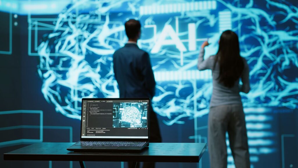 A laptop displaying code sits on a table in front of two people observing a large digital screen with neural network graphics and the letters "AI" illuminated, evoking an engaging video animation explainer atmosphere.