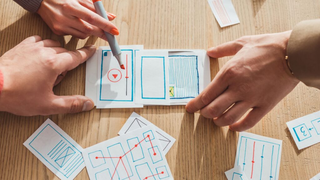 Two people sketch wireframes on paper for a mobile app interface, arranging layout and navigation elements alongside notes on animation best practices at a wooden table.