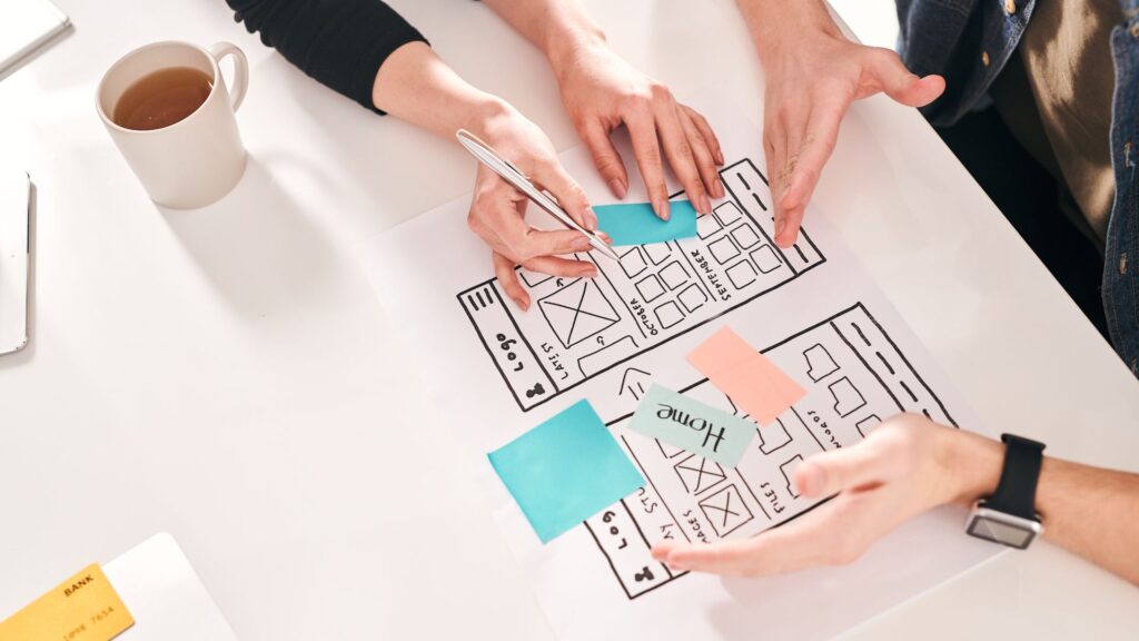 Three people collaborate on a website wireframe, adding and discussing sticky notes labeled with page names, while referencing animation best practices over a cup of coffee on the table.