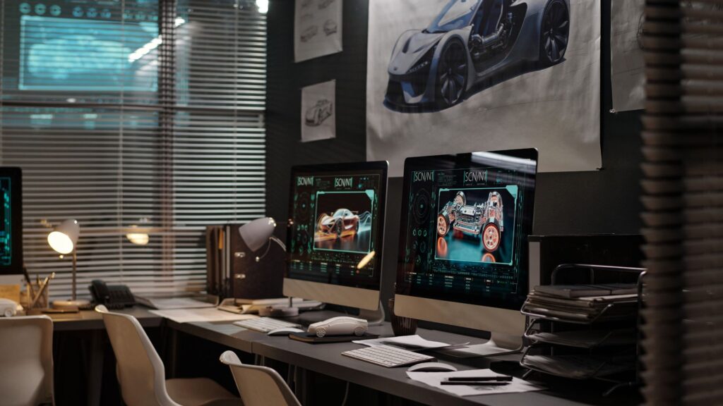 A modern office with computer workstations displaying car design software and animation elements, automotive sketches on the walls, and organized desks with office supplies.