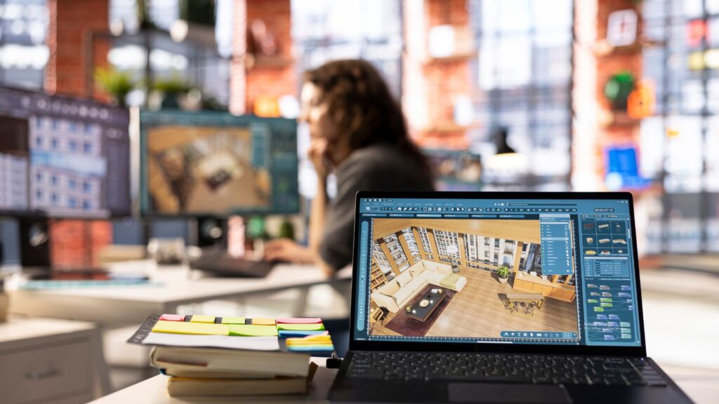 A laptop displays 3D interior design software with animation elements on a desk with notebooks and sticky notes; a person works at a computer in the blurred background.