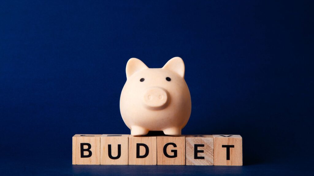 A piggy bank sits on wooden blocks that spell the word "BUDGET" against a dark blue background, highlighting smart savings with animation elements for added visual interest.