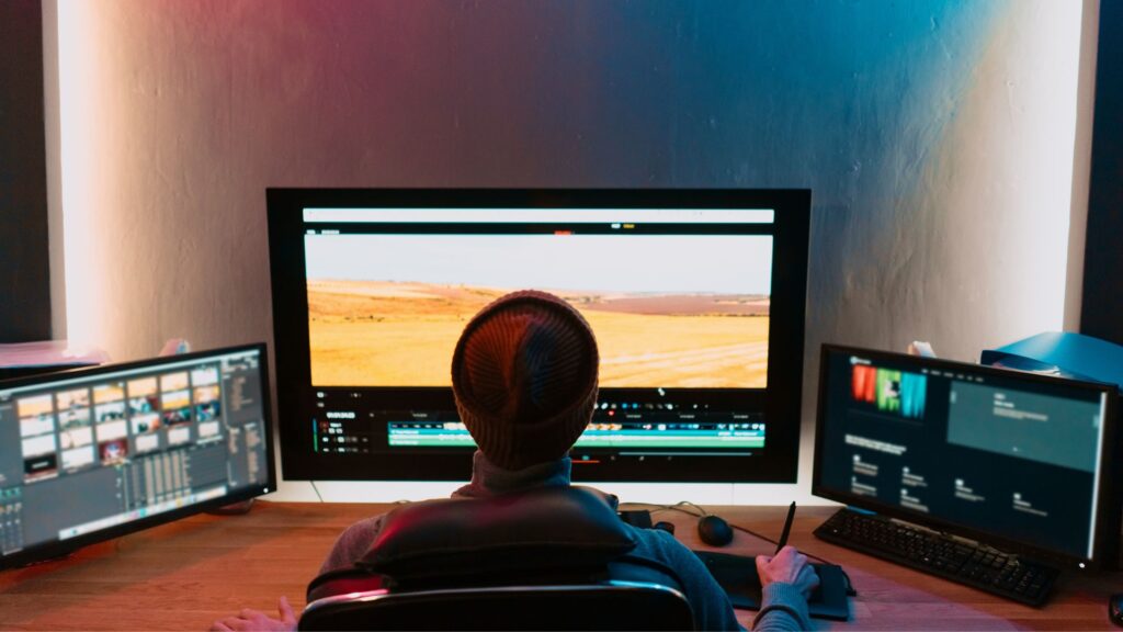 Person sitting at a desk with three monitors, editing a video animation on the central screen in a dimly lit room.