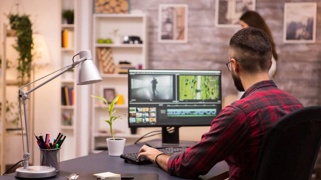 A man wearing glasses and a red plaid shirt edits video animation footage on a computer at a desk in a modern home office.