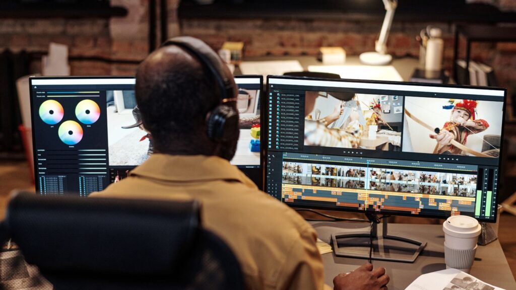 A person wearing headphones edits video footage and video animation on dual monitors at a desk with a keyboard, mouse, and a cup of coffee.