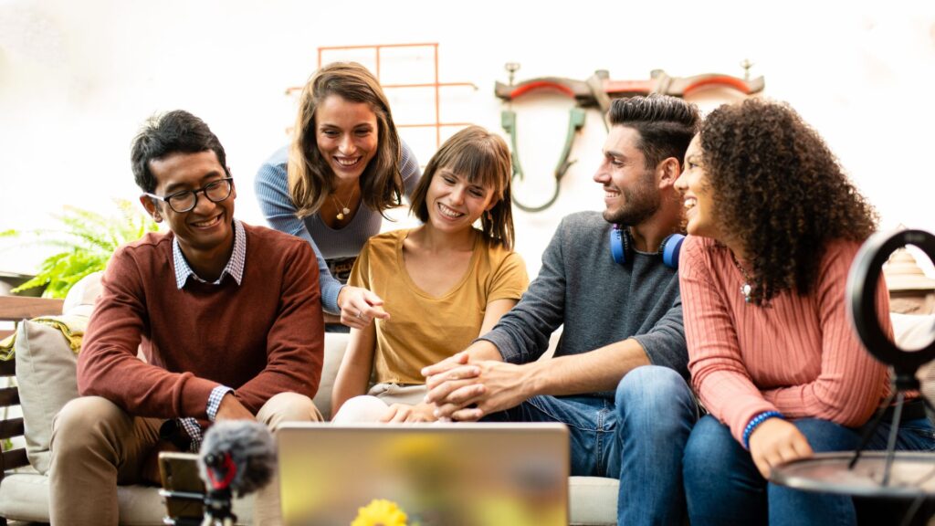 Five people sit together on a couch, smiling and looking at a laptop in front of them, appearing engaged in a group video animation activity or discussion.