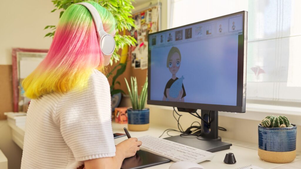 Person with multicolored hair and headphones uses a graphic tablet at a desk, working on a digital illustration for a video animation displayed on a computer monitor.