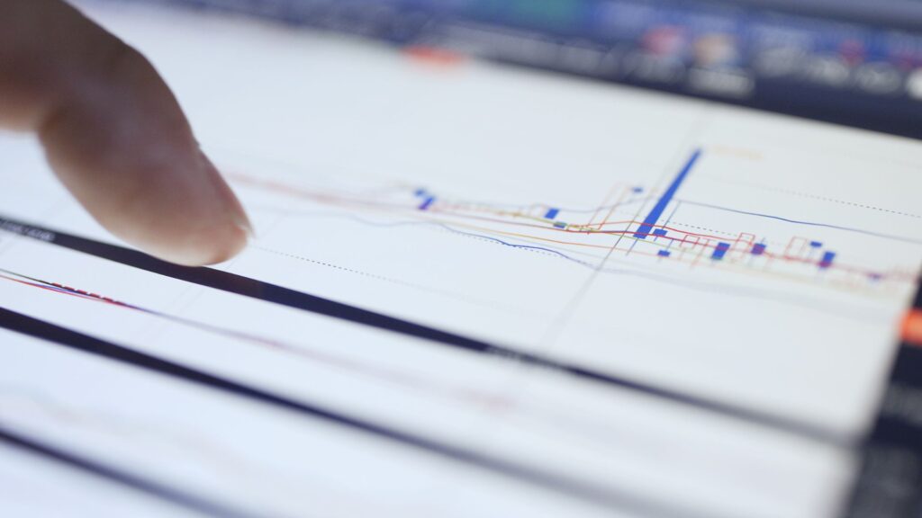A person points at a digital screen displaying a detailed financial chart with multiple lines and key metrics, emphasizing data-driven optimisation.