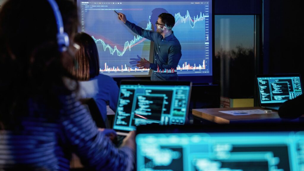 A person stands in front of a screen displaying key metrics and financial data, giving a presentation to an audience with laptops showing code, analytics, and optimisation strategies.