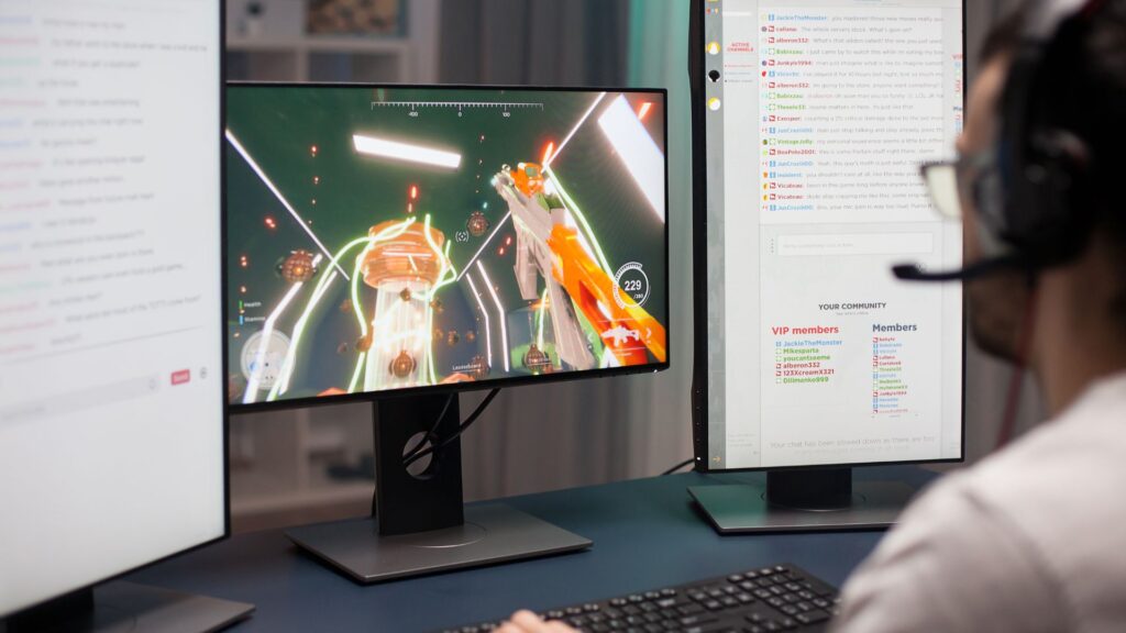 A person wearing a headset plays a sci-fi video game on a computer monitor while monitoring key metrics and live chat performance on a second screen.