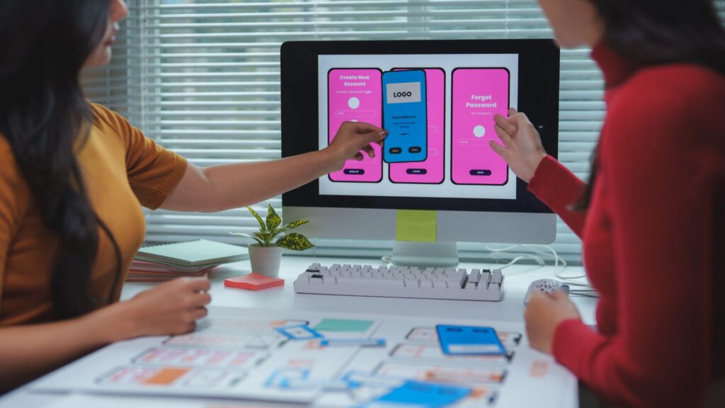 Two people discuss mobile app interface designs displayed on a computer monitor, pointing at screens showing create and reset password options. Design sketches, a phone, and notes on improving animation conversion rates are on the desk.