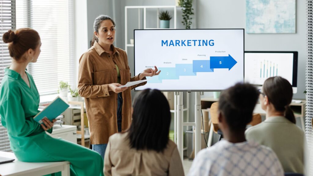 A woman presents an animation marketing strategy flowchart on a screen to a seated group in a modern office setting.