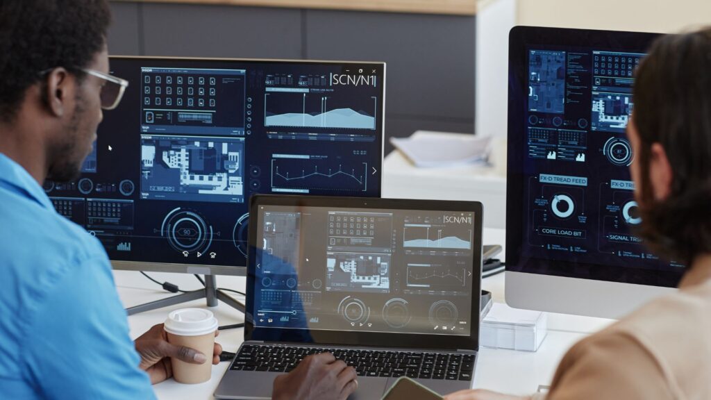 Two people sit at a desk working on computers displaying technical data, digital interface graphics, and an Animation Software Comparison; one holds a takeaway coffee cup.