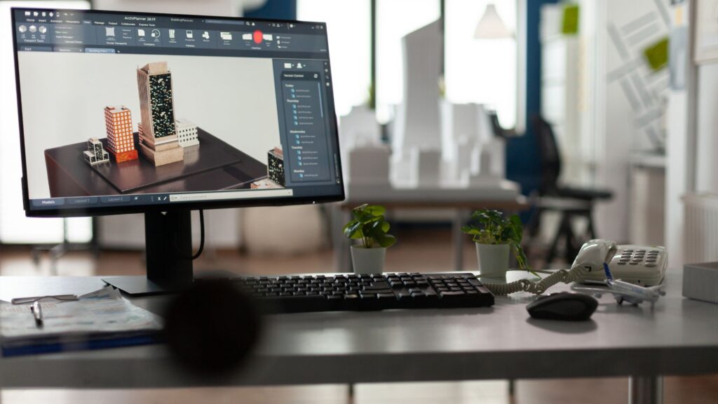 A computer monitor on a desk displays 3D architectural models in animation software comparison tools, with a keyboard, mouse, plants, and a phone nearby.