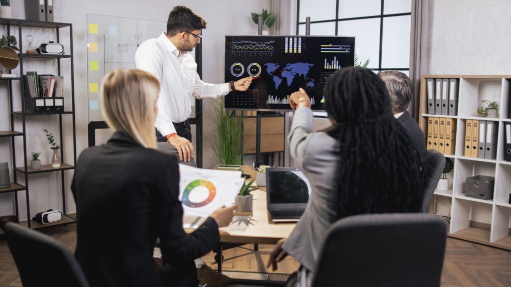 A man presents analytics charts and graphs on a large screen to three colleagues seated around a conference table in an office setting.