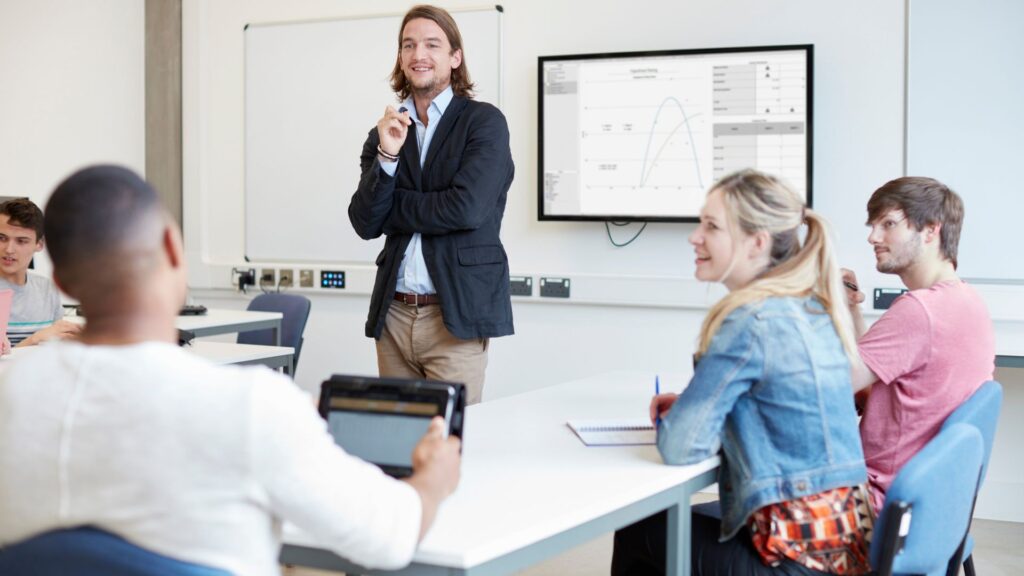 A man stands at the front of a classroom, providing practical guidance to three seated students; a graph is displayed on a screen behind him.