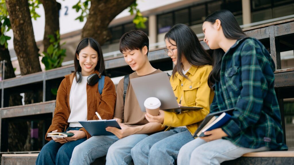 Four students sit outdoors on bleachers at an educational institution, looking at notes and a laptop together. One holds a clipboard, another a coffee cup, and two have books. Trees and a building are in the background—perfect for an educational animation scene.