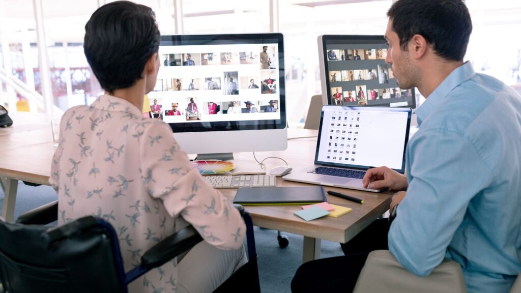 Two people sit at a desk viewing image thumbnails on large computer screens and a laptop in the bright office of a Startup Animation Company.