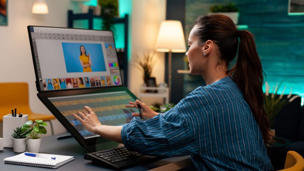 A woman uses a large touchscreen monitor and stylus to edit photos at her desk, with a keyboard, notebook, and plants nearby—perfect inspiration for those seeking Recommended Animation Services.